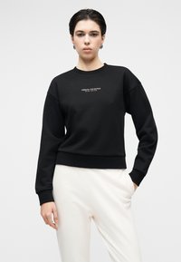 Μαύρο cropped φούτερ από υφασμάτινη ύφανση, με στρογγυλή λαιμόκοψη και ριμπ μανσέτες. Λευκό κεντημένο λογότυπο στο κέντρο.