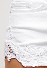 Primer plano de pantalones cortos de mezclilla blanca con un dobladillo de encaje floral, mostrando la textura de la tela y la costura de los bolsillos.