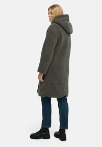 Grijze, fuzzy, capuchonjas met een gestructureerde afwerking, lange mouwen en zijsplitten, gedragen over donkerblauwe jeans en zwarte enkellaarsjes.