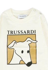 T-shirt in cotone color crema con una grafica a contorno nero di una testa di cane su uno sfondo quadrato dorato. L'etichetta riporta "TRUSSARDI JUNIOR."