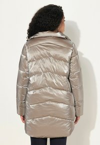 Metallisk pufferjacka med avtagbar huva, Chevron-quiltat mönster och långa ärmar. Färgen är ljus taupe med en slät textur.