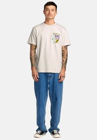 Lichtbeige katoenen T-shirt met een rechthoekige grafiek in paars, geel en groen op de linkerborst. Draagt loose blauwe jeans en zwarte sneakers.