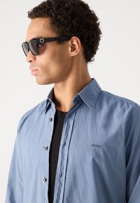Blue button-up shirt with a subtle texture and an embroidered "BOSS" logo, paired with dark sunglasses featuring thick black frames.