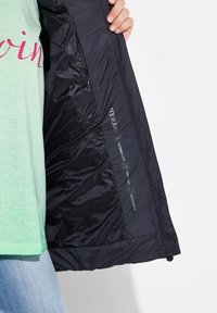 Schwarze wasserabweisende Jacke mit gequiltetem Innenfutter; zeigt Logos und Pflegezeichen. Darunter ist ein grünes Hemd zu sehen, getragen mit Jeans.