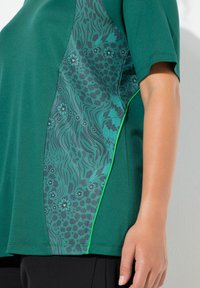 Zelený top s krátkými rukávy, který má texturovaný boční panel s motivy zvířat a květin v tmavších odstínech, doplněný světlým zeleným lemováním.