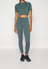 Completo crop top e leggings di colore verde scuro con cuciture di accento in turchese chiaro. Design aderente con texture liscia e maniche corte. Scarpe sportive nere.