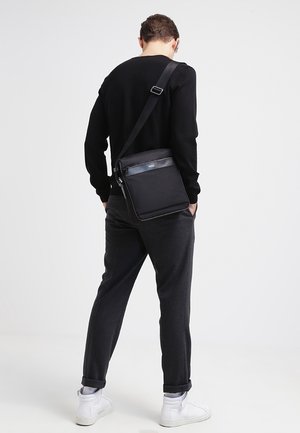 Schwarze Umhängetasche aus glattem Material, verstellbarer Riemen, vordere Reißverschlusstasche, quer getragen, kombiniert mit schwarzem Langarmshirt und dunkler Hose.