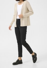 Beżowy kardigan z długim rękawem i otwartym przodem, połączony z czarnymi dżinsami slim fit i czarnymi mokasynami, idący na neutralnym tle.