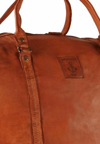 Sac de voyage en cuir marron présentant une texture lisse, des coutures détaillées, une forme rectangulaire et une étiquette en cuir estampillée avec un motif d'ancre.