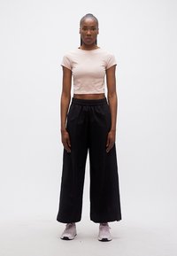 Γυναίκα που στέκεται προς τα εμπρός φορώντας ροζ ανοιχτό cropped t-shirt, μαύρο παντελόνι φαρδύ, και ροζ ανοιχτά sneakers σε απλό λευκό φόντο.