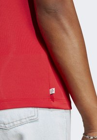 Czerwona bawełniana koszulka z gładką teksturą, posiada małą metkę z napisem "KONIEC Z ODPADAMI PLASTIKOWYMI", zestawiona z jasnoniebieskimi dżinsami.