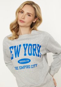 Šedá mikina s modrým nápisem "NEW YORK BROOKLYN THE EMPIRE CITY" v tučném písmu; měkká tkanina, klasický design s kulatým výstřihem.