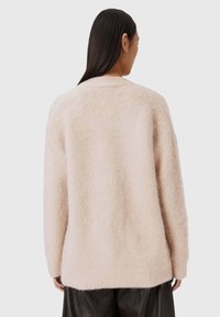 Kobieta z prostymi ciemnymi włosami, ubrana w luźny, jasnobeżowy, puszysty sweter oraz ciemne spodnie, odwrócona plecami do neutralnego tła.