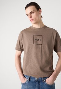 Brun bomulls T-skjorte med et firkantet logo som har teksten "BOSS" brodert i mørkebrunt. Kortermet og med rund hals, myk tekstur.