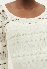 Haut en maille crochet écru, avec un encolure bateau, des motifs ajourés avec des boucles, et un tissu texturé léger.