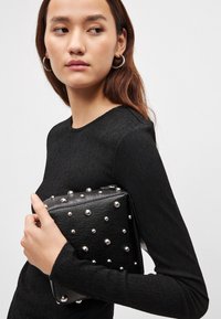 Schwarze Clutch mit strukturierter Oberfläche und silbernen Nieten, rechteckige Form, Reißverschluss, in der Hand gehalten. Model trägt ein schwarzes Langarmshirt.