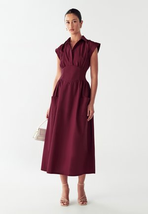 Abito bordeaux di lunghezza media con maniche corte, colletto, vita arricciata e due grandi tasche frontali. Tessuto liscio con un design strutturato.