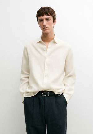 Jeune homme portant une chemise crème boutonnée rentrée dans un pantalon noir avec une ceinture noire, debout les mains dans les poches sur un fond blanc.