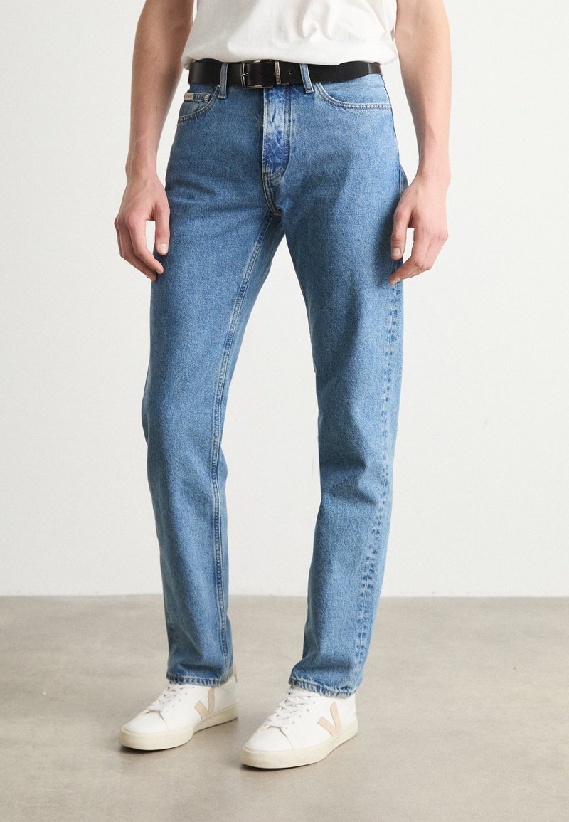 Jeans din denim bleu deschis, cu un croi drept, având cinci buzunare și o curea din piele neagră purtată la talie, asortate cu teneși albi.