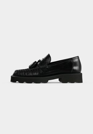 Mocassin en cuir noir avec détail pompon, couture avant texturée et semelle épaisse rainurée, présenté de profil sur fond blanc.