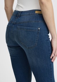 Vaqueros de mezclilla azul oscuro con cintura alta, que cuentan con bolsillos traseros y detalles de costuras sutiles. La pretina incluye una etiqueta color cuero.