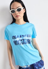 Blue cotton t-shirt with a graphic print text design. Round neckline, short sleeves. Model wears white oval sunglasses.