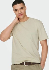 Beige Kurzarm-T-Shirt mit Rundhalsausschnitt. Hergestellt aus weichem Material, mit einem lässigen Schnitt. Trägt dazu eine khakifarbene Hose und einen braunen Gürtel.