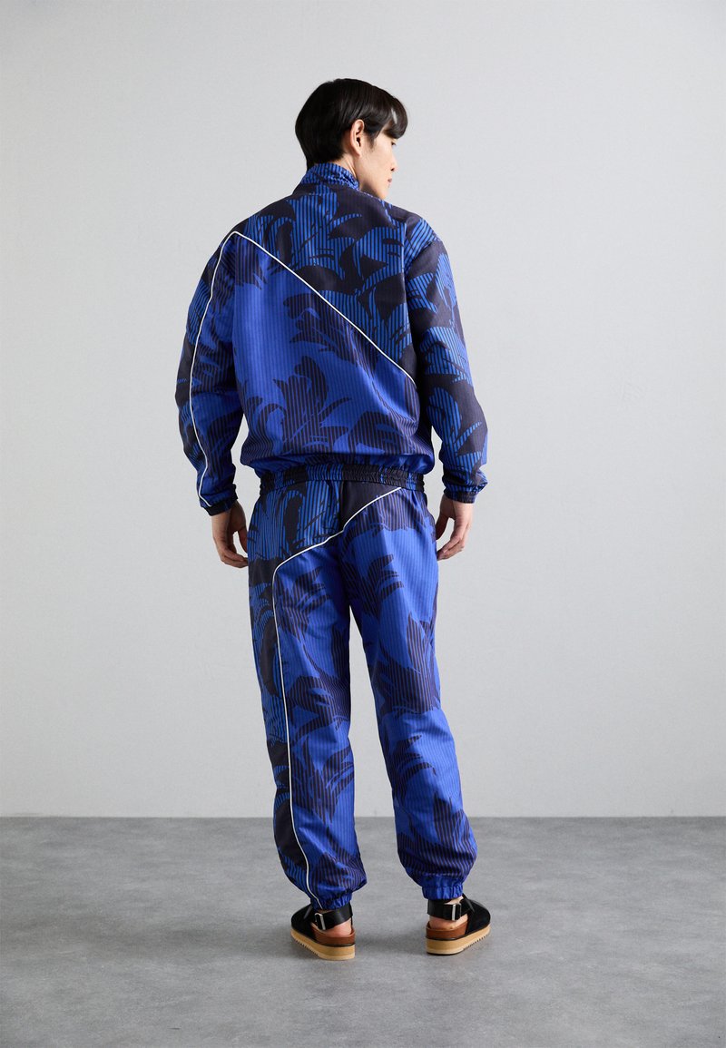 Blauwe trainingspak met grote zwarte bloemenpatronen en verticale strepen, inclusief een ritsjack en broek met elastische tailleband. Bruine sandalen.