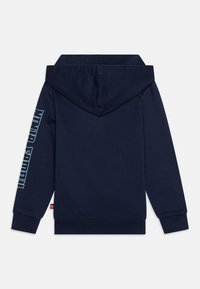 Mornarsko plava duks s kapuljačom, dugim rukavima i rebrastim manžetama. Veliki grafički natpis uz lijevi rukav. Mekana, glatka tekstura.