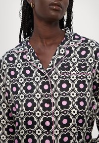 Haut de pyjama noir avec un motif floral rose et blanc, doté d'un col cranté, d'une poche et d'un passepoil rose contrastant le long des bords.