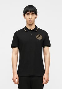 Άνδρας που φοράει μαύρο πόλο με γιακά και μανίκια διακοσμημένα με χρυσή λεπτομέρεια και λογότυπο Versace Jeans Couture σε χρυσό στη αριστερή πλευρά του στήθους.