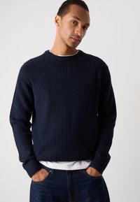 Navy ribbed knit sweater with a crew neckline and long sleeves. Textured fabric and relaxed fit. Worn over a white t-shirt and blue jeans.