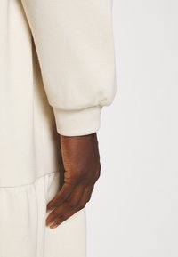 Sweatshirt beige clair avec une texture douce ; présente un poignet de manche froncé. Une main repose doucement sur le côté du vêtement.