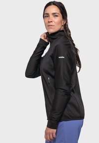 Schwarze Zip-Jacke mit hohem Kragen, langen Ärmeln, taillierter Passform und seitlichen Reißverschluss-Taschen. Markenlogo auf dem linken Arm sichtbar.