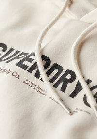 Felpa color crema con un grande logo nero "SUPERDRY". Spesso cordoncino con punte metalliche e tessuto morbido e testurizzato.