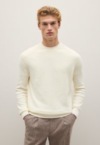 Cremfarbener gerippter Pullover mit rundem Ausschnitt und eng anliegenden Ärmeln, kombiniert mit braunen Herringbone-Muster-Hosen.