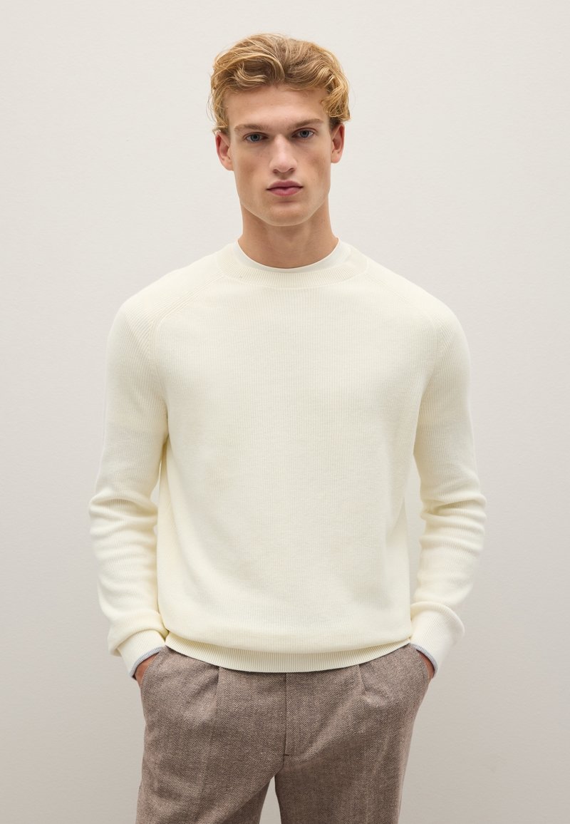Cremfarbener gerippter Pullover mit rundem Ausschnitt und eng anliegenden Ärmeln, kombiniert mit braunen Herringbone-Muster-Hosen.