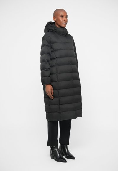 Črn puhovka, dolga in krojena, z brezšivno teksturo. Ima kapuco, visok ovratnik in stranske žepke. Obuta je z črnimi hlačami in gležnjački.