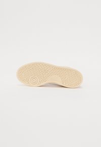 Semelle de basket de couleur claire avec une surface texturée et des crans proéminents, présentant un logo circulaire et un motif de bande de roulement complexe.