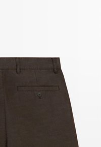 Vue arrière d'un pantalon marron foncé montrant la ceinture avec des passants et une poche arrière unique avec bouton sur fond uni.