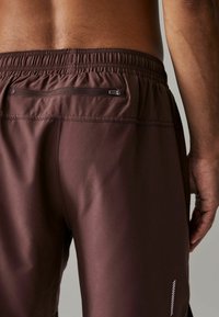 Brune atletiske shorts med elastisk talje, udstyret med en lille lynlås-lomme og hvide accentstriber nær kanten og taljen.