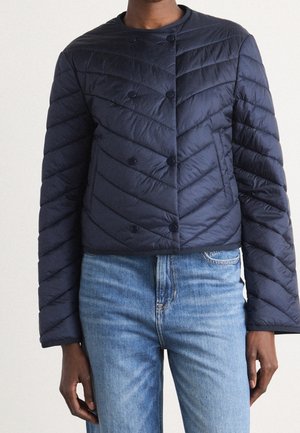 Person iført en mørkeblå quiltet jakke med trykknapper lagt sammen med lyseblå denimbukser mod en ensfarvet baggrund.