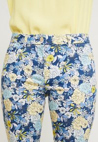 Blommigt mönstrade byxor med en blå bakgrund och gula samt ljusblå blommor. Tillverkade av tyg med en slät textur, inklusive en framknapp.