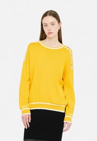 Maglione giallo a maglia con accenti bianchi, colletto ampio a giro e dettagli con bottoni laterali. Modello indossa una gonna nera. Tessuto lavorato, vestibilità comoda.