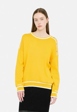 Maglione giallo a maglia con accenti bianchi, colletto ampio a giro e dettagli con bottoni laterali. Modello indossa una gonna nera. Tessuto lavorato, vestibilità comoda.