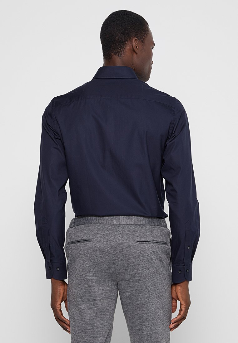 Mężczyzna w granatowej koszuli z długim rękawem, włożonej w szare, wzorzyste spodnie, stoi tyłem do jednolitego tła.