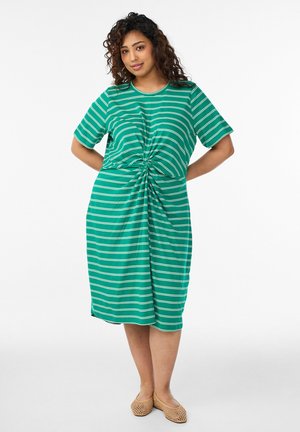 Žena v zeleno-bielych pruhovaných šatách po kolená s krátkymi rukávmi a detailom prekrútenia vpredu, stojaca s rukami za chrbtom.
