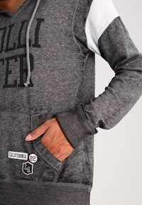 Šedá mikina s kapucí s kontrastními bílými rukávy, velkým černým nápisem na přední straně a kapesou, na které je nápis "CALIFORNIA" a červené logo.