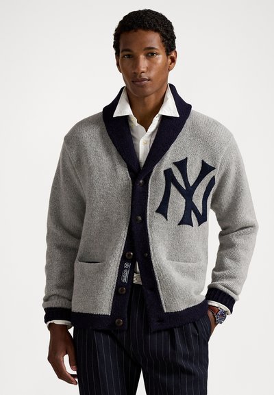 Homme portant un cardigan à col châle gris avec des finitions bleu marine, un grand logo NY, une chemise blanche à boutons, un pantalon à rayures et une montre au poignet, les mains détendues.