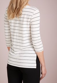 Frau in einem weißen Langarmshirt mit dünnen schwarzen horizontalen Streifen und schwarzen Hosen, die vor einem hellrosa Hintergrund steht.